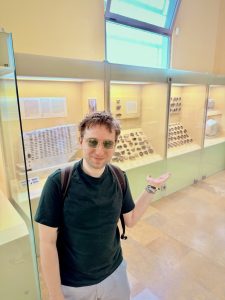 Ich stehe in einem Museum vor Tonscherben die mit Namen beschriftet sind (Ostrakon) und einer antiken Losmaschine zur Besätzung von Ämtern (Kleroterion)